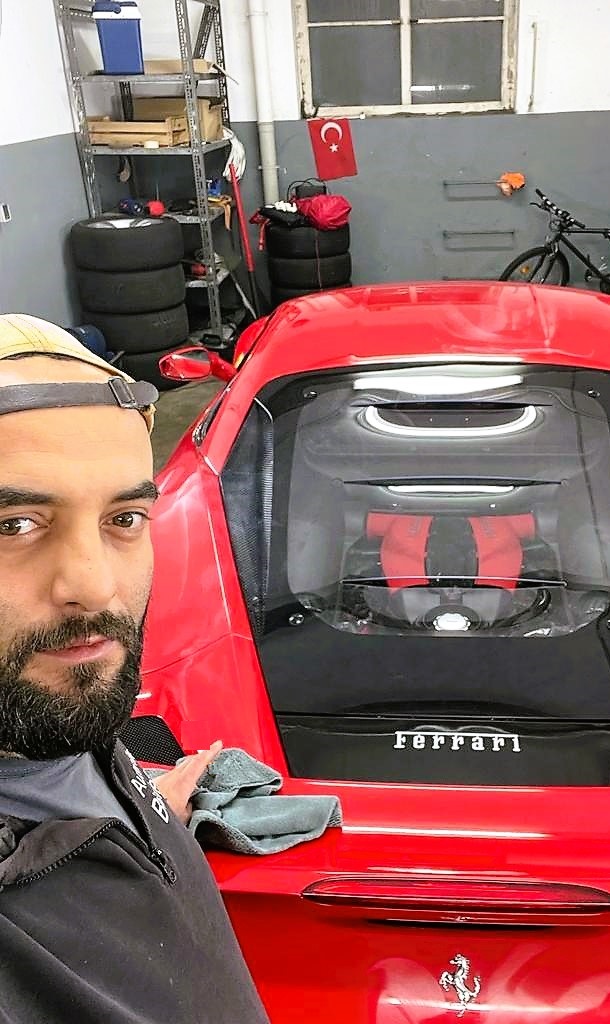 Mann mit Bart und Mütze lächelt leicht, steht neben rotem Ferrari in Werkstatt mit Reifenregalen.