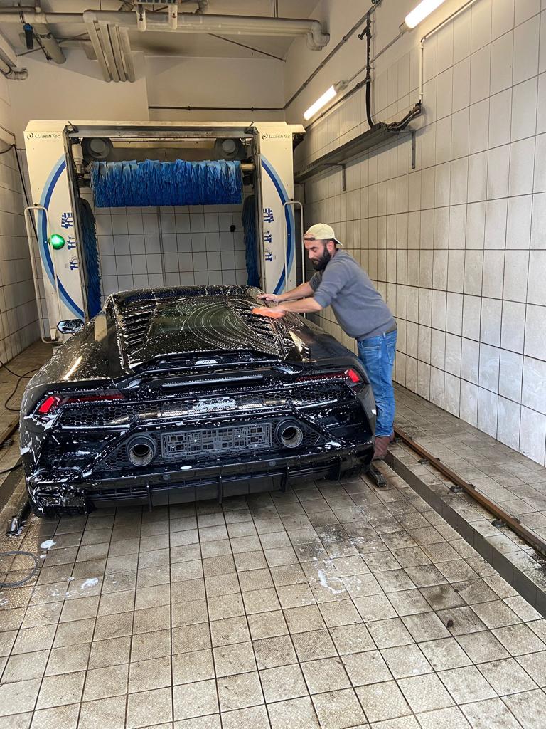 Mann wäscht schwarzen Sportwagen in Waschanlage, lächelt, trägt Baseballmütze und graues Oberteil.