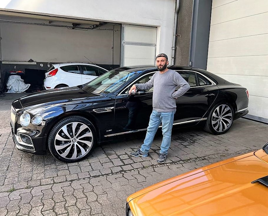 Mann mit Bart lächelt, steht neben schwarzen Luxusauto mit großen Rädern, trägt grauen Pullover.