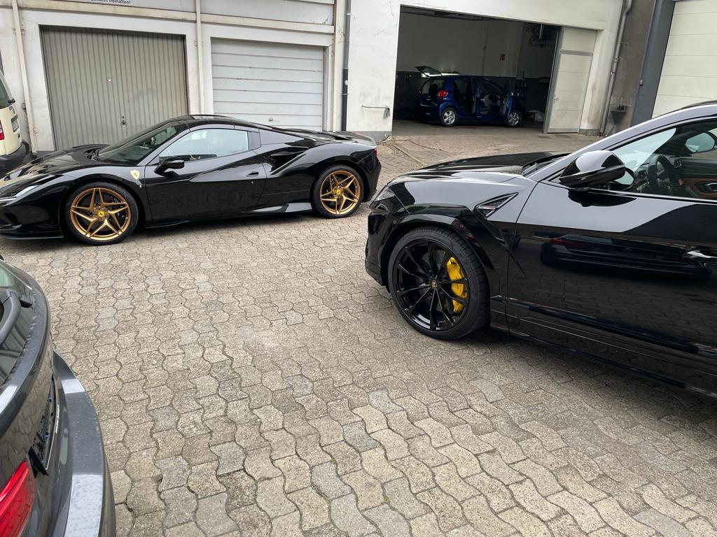 Zwei schwarze Sportwagen mit goldenen Felgen auf gepflastertem Hof geparkt.